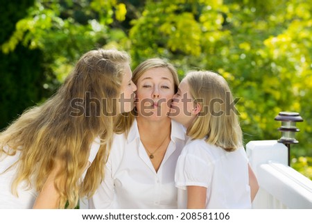 Front view of mother being kissed by her two daughters outdoors on patio with woods in background 