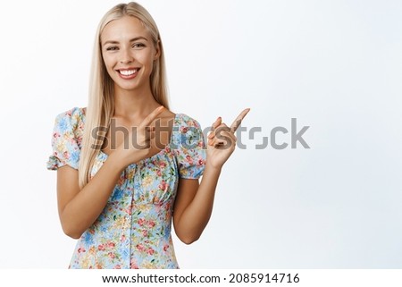 Cute blond girl smiles, points at upper right corner, shows advertisement, announcement or logo, stands against white background