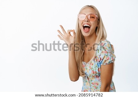 Beautiful and stylish blond woman in sunglasses, laughing and smiling, showing okay ok gesture, standing in dress over white background