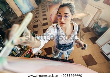 Artist painting while wearing tinted eyeglasses. Imaginative young painter making a new artwork for her project. Creative female artist standing in front of a canvas in her atelier. Royalty-Free Stock Photo #2085949990