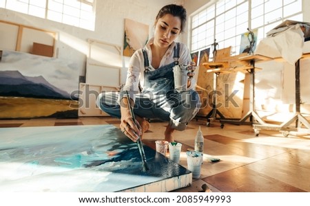 Female artist painting on a large canvas with a paintbrush. Imaginative young woman working on the floor of her art studio. Creative female painter making a blue artwork for her new project. Royalty-Free Stock Photo #2085949993