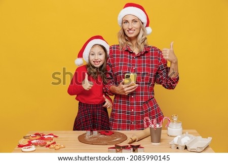 Two woman mother, fun child baby girl in red Christmas hat isolated on plain yellow wall background studio. Mom little kid cook ginger cookie at kitchen table home. Happy New Year 2022 holiday concept