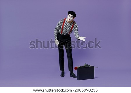Full size body length young mime man with white face mask wears striped shirt beret show point hand down on suitcase bag rose flower isolated on plain pastel light violet background studio portrait