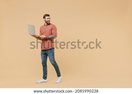 Full size body length joyful tattooed young brunet man 20s short haircut wears apricot shirt go stride hold use laptop pc computer look back behind isolated on pastel orange background studio portrait