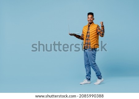 Full size body length young black man 20s years old wears yellow waistcoat shirt hold use work on laptop pc computer doing winner gesture isolated on plain pastel light blue background studio portrait