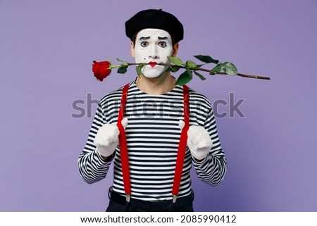 Full size body length romantic young mime man with white face mask wears striped shirt beret hold in mouth rose flower looking camera isolated on plain pastel light violet background studio portrait
