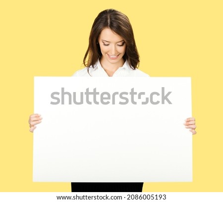 Portrait image of cheerful smiling woman in white confident wear holding showing mock up empty signboard. Businesswoman in advertising studio concept. Copy space area. Yellow color background. Advert