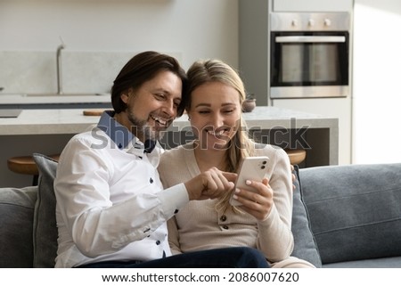Happy bonding 35s family couple using cellphone software applications, choosing goods in internet store, watching funny photo video content in social network, modern technology addiction concept.