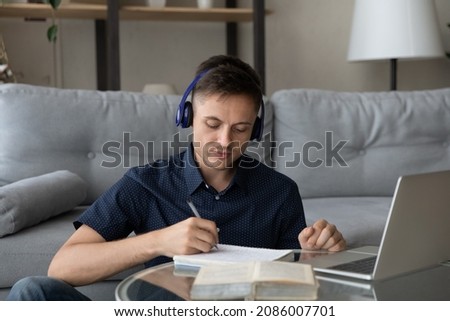 Focused young adult student guy wearing wireless earphones, making notes, writing in notebook, listening to online learning lecture, music for study, deep focus, concentration, working at laptop