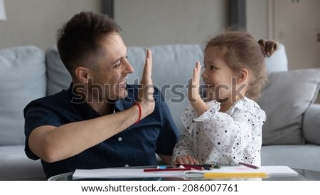 Happy excited dad and daughter girl giving high five over album with drawings, colorful pencils. Father showing praise, support for good homework result, playing creative learning games with child