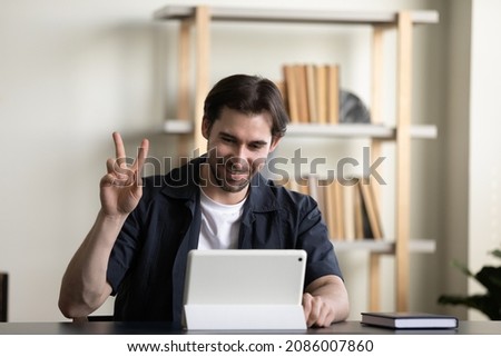 Positive millennial employee guy, tablet user waving hello at screen, showing peace, victory hand finger gesture, making distant video call to friend, coworker, client at workplace table