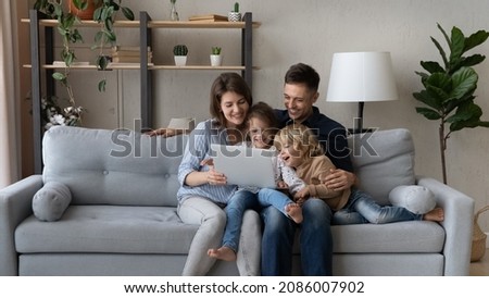 Cheerful millennial parents resting with gen Z kids on couch, making video call, watching movie online, internet TV on laptop computer, having fun, laughing. Happy family enjoying weekend