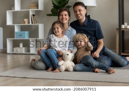 Happy sweet family posing on photo shooting at home, sitting on warm heating floor together, looking at camera away, smiling, laughing. Millennial couple of parents hugging sibling kids. Home portrait