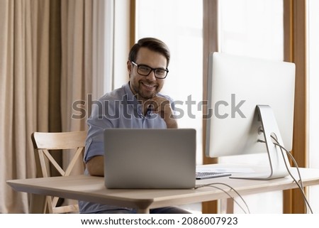Happy confident professional man working at laptop and desktop, two computers, sitting at workplace, smiling, looking at camera. Business man, programmed, financial trader business head shot portrait