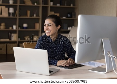 Happy millennial Indian employee satisfied with job, working at laptop, workstation monitor, typing, looking away, laughing. Young business woman, professional, marketing finance analyst at workplace