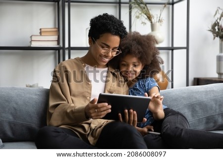 Happy bonding young African American mother or babysitter in eyeglasses using digital computer tablet with laughing adorable small kid girl, web surfing, shopping online or playing games at home.