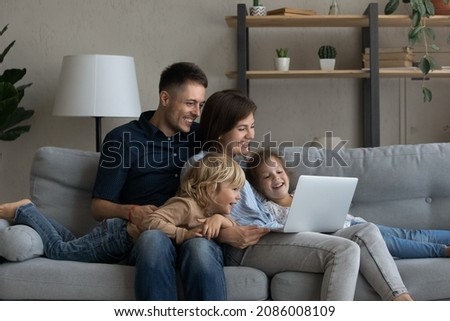 Happy joyful family watching online movie on laptop, relaxing on sofa together, enjoying internet TV channel, making video call. Millennial parents and two children resting at home
