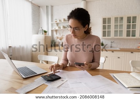 Confident woman in glasses calculating family budget, using smartphone and calculator, internet banking service, working with financial documents in kitchen, paying bills online, managing expenses Royalty-Free Stock Photo #2086008148
