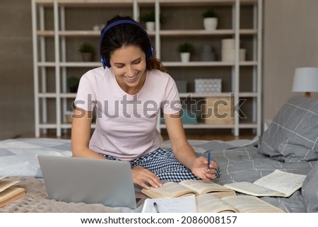 Smiling woman in wireless headphones using laptop, studying online at home, positive young female student watching webinar listening to lecture, writing taking notes, sitting on bed with books
