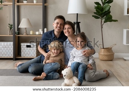 Happy parents and two little sibling kids home portrait. Millennial family couple hugging preschool children, sitting, playing on floor at home, looking at camera, smiling, laughing