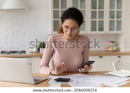 Confident woman in glasses counting bills, calculating budget at home, using calculator and smartphone apps, working with financial documents in kitchen, paying bills online, managing expenses Royalty-Free Stock Photo #2086008256