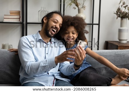 Happy laughing young African American father and little adorable kid daughter looking at cellphone screen, watching funny video in social network, playing mobile games, modern tech addiction concept.