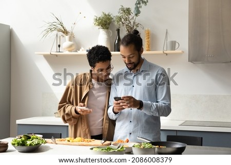 Distracted from cooking happy young African American family couple using cellphones, checking interesting food recipe, communicating online, inviting friends for party, preparing meal in kitchen.