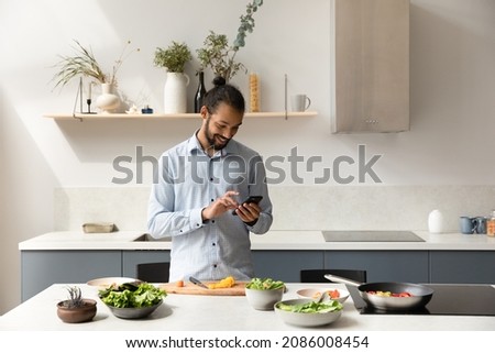 Happy young African American man using cellphone, searching interesting food recipe online, communicating in social network, cooking healthy meal alone in modern kitchen, modern tech addiction.
