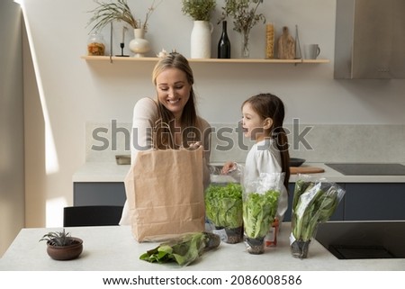 Happy young mother and joyful cute small kid daughter unpacking fresh salad leaves and organic greenery from carton package, unpacking products for healthcare eating together in modern kitchen.