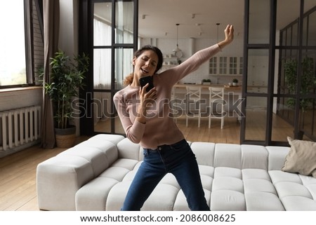 Overjoyed funny woman holding smartphone, having fun in modern living room, happy young woman excited by good news, dancing to music on phone, spending leisure time with modern device at home