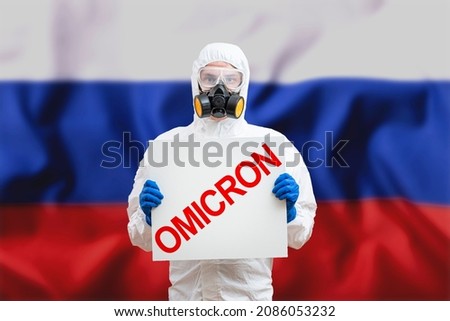 Medical worker wearing PPE protective white suit,face shield mask, holding text inscription omicron covid-19 SARS-CoV-2 Coronavirus B.1.1.529 new variant or strain behind blur Russia flag
