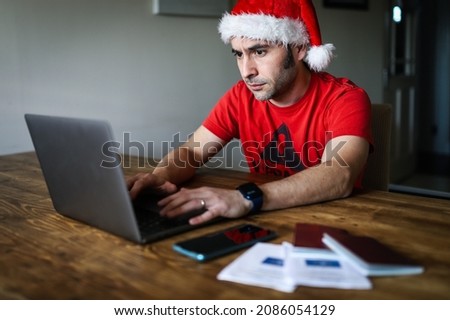 A man with a santa hat using a laptop with passports, a cellphone and covid digital certificates lying on the table out of focus - booking tickets during christmas concept