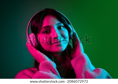 Music at headphones. Young adorable cute girl, student in casual style clothes posing isolated on dark green studio backgroud in pink neon light. Emotions, facial expression, youth, fashion and ad
