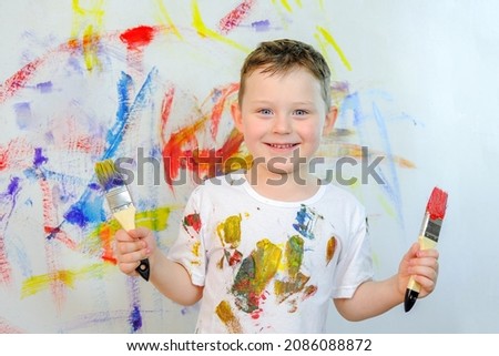 A handsome smiling blue-eyed boy stands against the background of a colorful wall. The child holds a brush in his hands. Boy in a white T-shirt, stained with paint. Wall in colored paints. Royalty-Free Stock Photo #2086088872
