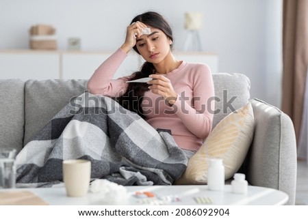 Despaired sad young hindu lady with plaid, suffer from flu, sits on sofa and looks at thermometer in living room interior. Cold, fever, disease treatment at home and covid-19 lockdown, free space Royalty-Free Stock Photo #2086092904