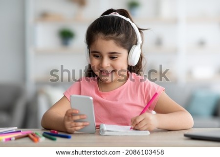 Remote Education. Smiling Little Girl In Headphones Using Smartphone At Home For Distance Learning, Cute Arab Female Kid Sitting At Desk, Looking At Cellphone Screen And Writing In Notepad, Closeup