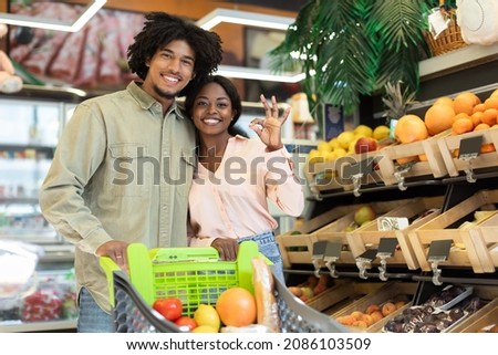 Happy Black Couple Gesturing Okay Doing Grocery Shopping Posing Standing With Shop Cart Full Of Food Products, Smiling To Camera In Modern Supermarket Indoors. We Are OK