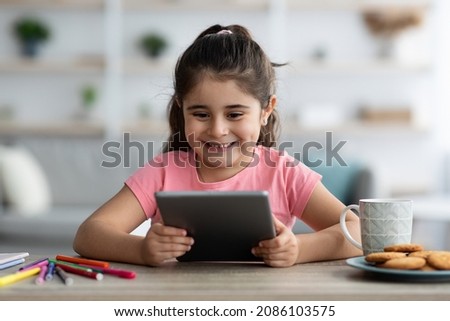Gadgets For Kids. Happy Little Arab Girl Relaxing With Digital Tablet At Home, Cute Female Child Sitting At Desk In Living Room And Watching Cartoons On Pad Computer, Closeup Shot With Copy Space