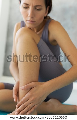 Attractive girl sitting on floor, stroking hurt leg after training session at fitness studio, dressed in violet crop top and leggings, having sprain or cramp in result of hard workout