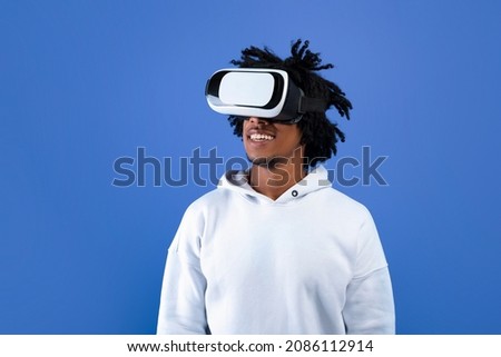 Cool African American teen guy wearing VR headset, using virtual reality for entertainment on blue studio background, copy space. Funky black youth exploring cyberspace. Futuristic technologies