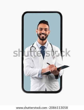Medical Services. Young Happy Male Middle Eastern Doctor In White Uniform With Stethoscope And Clipboard Writing Prescription To Patient And Smiling At Camera, Looking Coming Out Big Cell Phone Screen Royalty-Free Stock Photo #2086113058