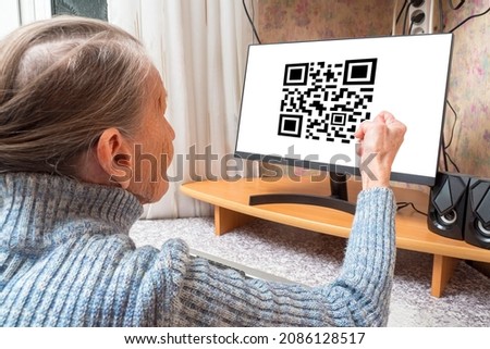 Angry elderly woman shakes her fist at a white computer screen with a qr code. Aggressive emotions of the grandmother in front of the TV. Сoncept is a protest against forced vaccination or isolation.