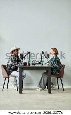 Vertical full length portrait of two people speaking to microphones while recording podcast in studio