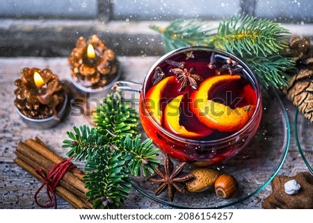 Glass of mulled wine, punch, hot tea, with orange slices with cinnamon sticks in beautiful Christmas atmosphere on rustic wood in front of old wooden window in winter night landscape.
