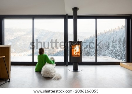 Woman sitting with dog near fireplace and panoramic window at modern living room with stunning view on snowy mountains. Concept of rest in houses or cabins on nature. Idea of escape from everyday life Royalty-Free Stock Photo #2086166053