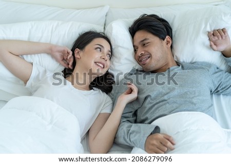 Portrait of Asian new marriage couple lying on bed and look each other. Attractive beautiful young man and woman in pajamas enjoy early morning activity in bedroom at home. Family relationship concept