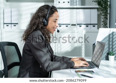 Latino beautiful business woman call center smile while work in office. Attractive young female employee worker in formal wear sit on table at workplace, use laptop computer talk to support customer.