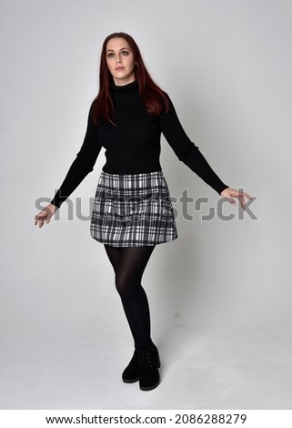 Full length portrait of redhead girl wearing black autumn fashion with plaid skirt and boots. Standing pose with hand gesture  isolated on  studio background 