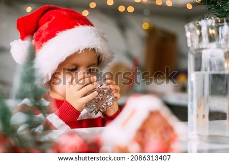 Happy funny toddler boy in a red Santa hat drinking filtered water from a glass in the kitchen. Holidays, health concept.