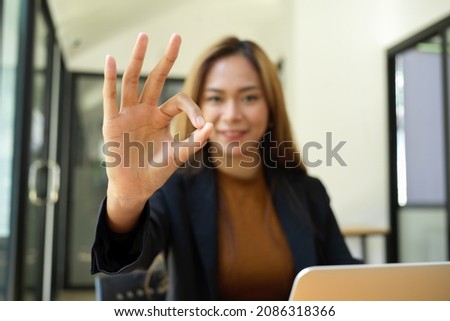 Cheerful asian young business worker posting OK hand sign at her desk. deal, agreement, happy, ok gesture.